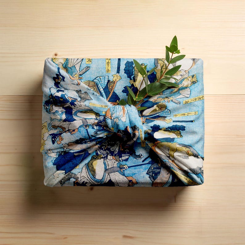 May include: A square package wrapped in blue fabric with a Japanese illustration, tied with a knot. The fabric depicts figures in blue, white, and tan. A sprig of green leaves is tucked into the knot.