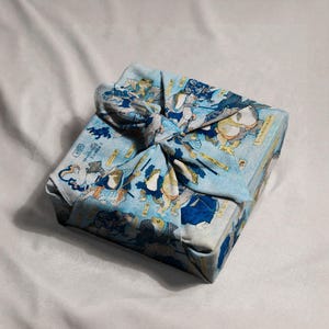 May include: A square package wrapped in a light blue cloth with a Japanese-style print featuring illustrations of frogs in traditional attire. The cloth is tied in a decorative knot on top, creating a unique presentation.