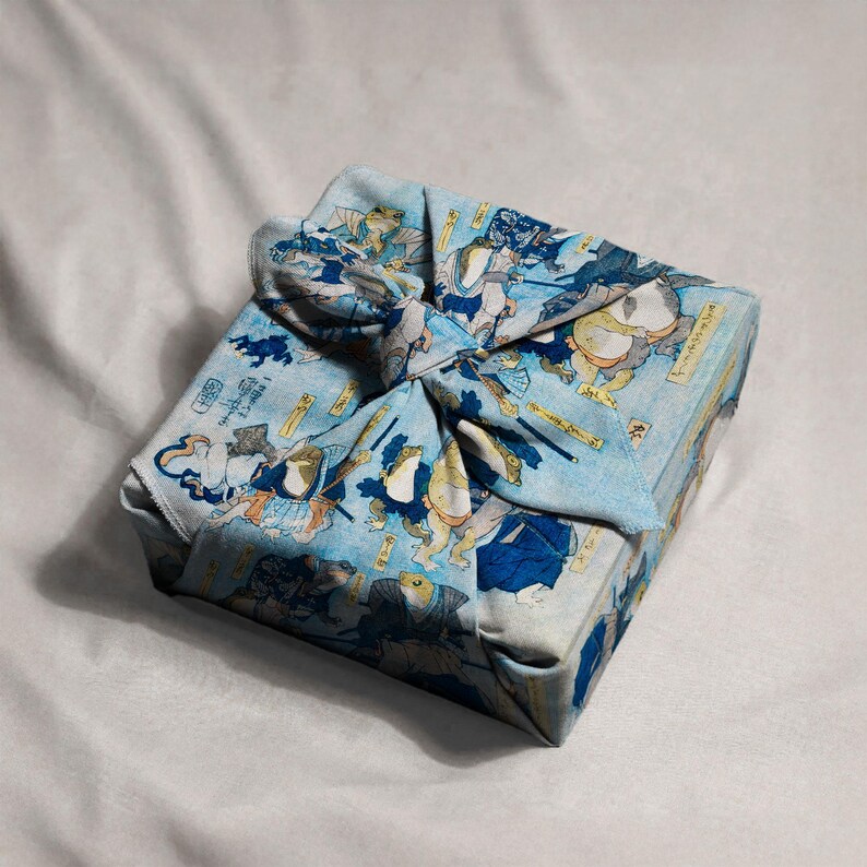 May include: A square package wrapped in a light blue cloth with a Japanese-style print featuring illustrations of frogs in traditional attire. The cloth is tied in a decorative knot on top, creating a unique presentation.