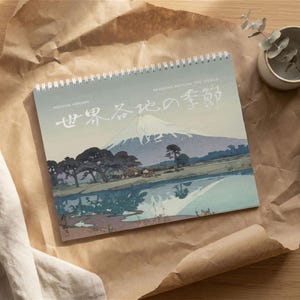 May include: A spiral-bound wall calendar featuring a landscape print of Mount Fuji with a reflective lake, dark green trees, and small buildings. The text reads Yoshida Hiroshi and Seasons Around the World in English and Japanese characters.