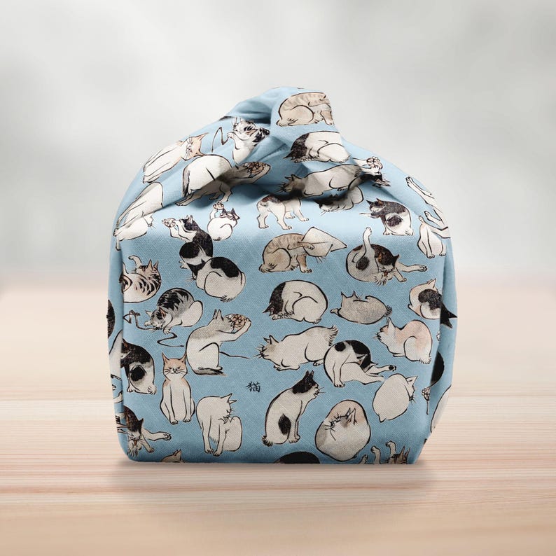 Pu&ograve; includere: Una borsa di tessuto azzurro chiaro con un motivo ripetuto di gatti bianchi e neri in varie pose. La borsa &egrave; annodata in alto, creando una forma arrotondata. La parola "猫" &egrave; stampata sul tessuto.