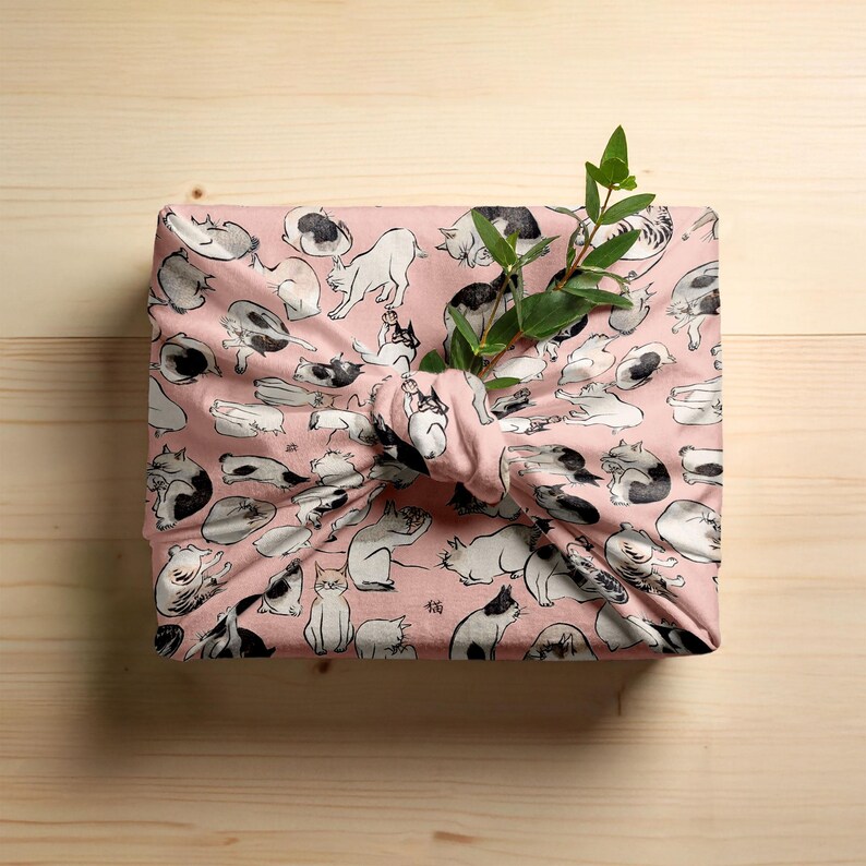 Pu&ograve; includere: Un involucro di tessuto rosa con un motivo ripetuto di illustrazioni di gatti in bianco e nero. Il tessuto &egrave; annodato e un rametto di foglie verdi &egrave; infilato nel nodo. L'involucro &egrave; su una superficie di legno chiaro.