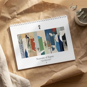 May include: A spiral-bound calendar titled "Seasons of Japan" featuring twelve curated artworks. The calendar displays colorful, vertical panels with Japanese-inspired illustrations. The calendar is on a brown paper surface with a small plant in a ceramic pot.