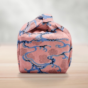 Pu&ograve; includere: Una borsa in tessuto rosa con un motivo a onde blu e una parte superiore annodata. La borsa presenta un motivo ripetuto di onde blu e forme simili a nuvole su uno sfondo rosa. La borsa &egrave; appoggiata su una superficie di legno.
