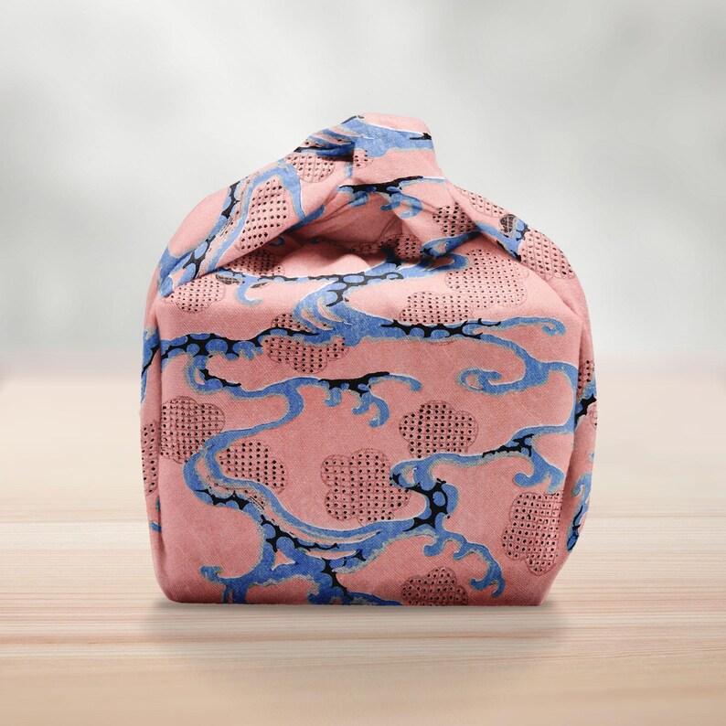 Pu&ograve; includere: Una borsa in tessuto rosa con un motivo a onde blu e una parte superiore annodata. La borsa presenta un motivo ripetuto di onde blu e forme simili a nuvole su uno sfondo rosa. La borsa &egrave; appoggiata su una superficie di legno.