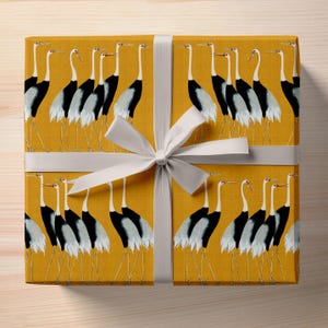 May include: Gift-wrapped present featuring a mustard-yellow paper with a repeating pattern of stylized cranes in black, white, and gray. The gift is tied with a wide, light gray satin ribbon and bow, creating a visually appealing package.