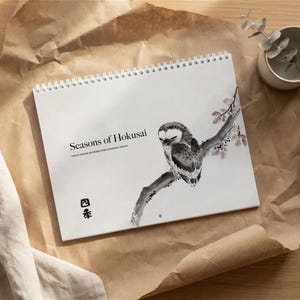 May include: A spiral-bound calendar titled "Seasons of Hokusai" featuring a black and white illustration of an owl perched on a branch with pink blossoms. The calendar is on a brown paper surface.