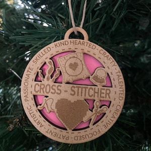 May include: A round wooden ornament with a pink background and the words "CROSS-STITCHER" in the center. The ornament is decorated with images of cross-stitch supplies and the words "PATIENT - ARTISTIC - FOCUSED - CREATIVE - TALENTED - CRAFTY - KIND HEARTED - SKILLED".
