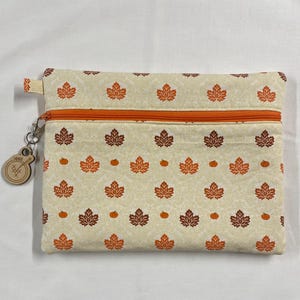 May include: A cream-coloured zippered pouch featuring a pattern of autumnal maple leaves and pumpkins in shades of orange and brown. The pouch has an orange zip and a wooden charm attached to the zip pull.