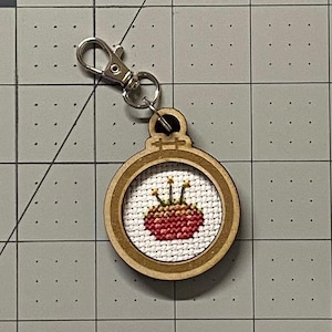 May include: A small wooden framed embroidery hoop keychain with a red and yellow embroidered strawberry design.