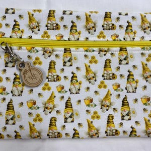 May include: A white zippered pouch with a repeating pattern of yellow and black bee-themed gnomes, honeycombs, and bees. The pouch has a yellow zipper and a wooden charm with a metal clasp.