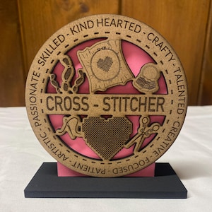 May include: A wooden plaque with a pink background and a cutout design of a cross-stitch project, scissors, and a needle. The plaque is inscribed with the words "CROSS-STITCHER" and a list of positive attributes, including "skilled", "kind hearted", "crafty", "talented", "creative", "focused", "patient", and "artistic".