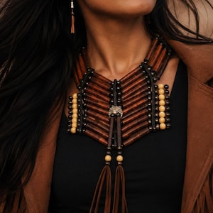 Può includere: Una collana importante con tubi in legno marrone scuro disposti orizzontalmente, piccole perline color crema e distanziatori metallici scuri. Il pezzo presenta un accento centrale circolare in metallo con due lunghe nappe in camoscio marrone.