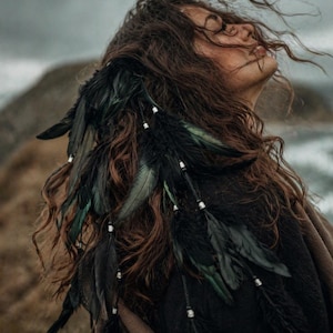Può includere: Un accessorio per capelli con piume nere e fili di perline bianche. L'accessorio è attaccato a lunghi capelli castani ondulati. Lo sfondo è una scena all'aperto sfocata con un cielo nuvoloso.