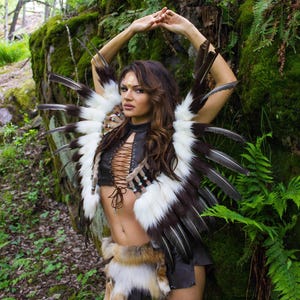 May include: A woman wearing a brown leather and white fur vest with black feathers. The vest has a lace-up front and is decorated with beads. She is also wearing a brown leather skirt with a fur trim.