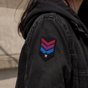 May include: A black denim jacket featuring a shield-shaped patch on the sleeve. The patch displays three horizontal chevron stripes in magenta, purple, and blue, with a small white star centered at the bottom point against a black background.