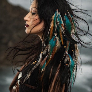 May include: A woman with long black hair adorned with colorful feathers. The feathers are in shades of teal, brown, and tan, and are attached with small beads. The woman is wearing a dark top and a brown shawl.