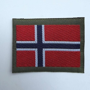 Puede incluir: Un parche rectangular bordado que presenta la bandera de Noruega con fondo rojo, una cruz azul oscuro y bordes blancos. El diseño de la bandera está rodeado por un borde grueso de color verde oliva con una textura de tejido visible.