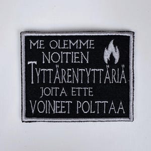 May include: A black rectangular patch with white embroidered text and a flame graphic. The text reads "ME OLEMME NOITIEN TYTTÄRENTYTTÄRIÄ JOITA ETTE VOINEET POLTTAA". The patch has a white border.