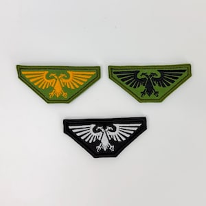 Op de afbeelding: Drie trapeziumvormige geborduurde patches met een tweekoppig adelaarontwerp. Eén patch heeft een groene achtergrond met een gele adelaar, één een groene achtergrond met een zwarte adelaar en één een zwarte achtergrond met een witte adelaar.