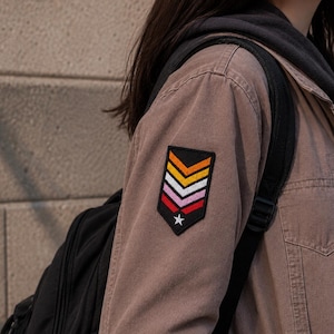 May include: A tan jacket featuring a shield-shaped embroidered patch on the sleeve. The patch has a black background with four horizontal chevron stripes in orange, yellow, white, and pink, finished with a small white star at the bottom point.