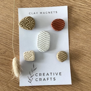 May include: Set of five clay magnets in various shapes and colors, including brown, beige, white, and red. The magnets have a textured surface and are arranged on a white card with the text "CLAY MAGNETS" and "CREATIVE CRAFTS".