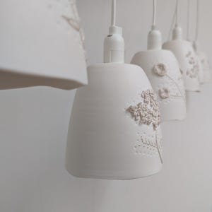 Op de afbeelding: Een rij witte keramische hanglampen met bloemreliëfdetails. Elke lamp heeft een belvorm en wordt opgehangen aan een wit snoer. De bloemmotieven zijn in een lichtere tint, wat textuur en visuele interesse aan de lampen toevoegt.