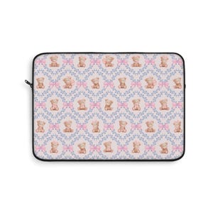 May include: A laptop sleeve featuring a repeating pattern of teddy bears, pink bows, and blue floral elements on a light pink background. The sleeve has a black zipper and trim, offering a decorative and protective design.