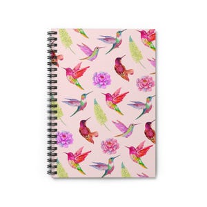 Può includere: Quaderno a spirale con un motivo ripetuto di colibrì colorati, fiori rosa e fogliame verde su sfondo rosa chiaro. Il quaderno ha una rilegatura a spirale nera e una copertina bianca.