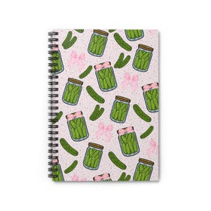 Op de afbeelding: Spiraalgebonden notitieboek met een herhalend patroon van cartoon potten augurken, groene augurken en roze strikken op een lichtroze gespikkelde achtergrond. Het notitieboek heeft een zwarte spiraalbinding.