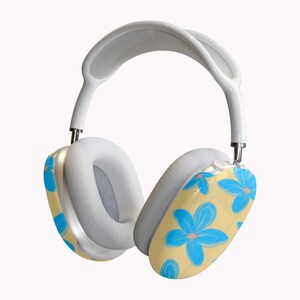 May include: White over-ear headphones with a floral design. The earcups have a light yellow background with blue flowers and pink centers. The headband and ear cushions are white.