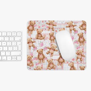 Peut inclure: Tapis de souris rectangulaire avec un motif répété d'ours en peluche avec des nœuds roses sur un fond floral et à carreaux. Une souris d'ordinateur blanche repose sur le tapis, à côté d'un clavier blanc.