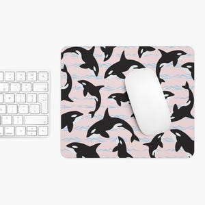 May include: A rectangular mousepad with a pink background featuring a pattern of black and white orcas and light blue wave lines. A white computer mouse rests on the pad. A white keyboard is visible on the left.