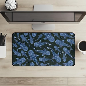 May include: A rectangular mousepad featuring a pattern of blue whale sharks on a dark green background. The whale sharks have white spots and are arranged in various directions. The mousepad is on a light wood surface, with a computer monitor, coffee cup, and other desk accessories.