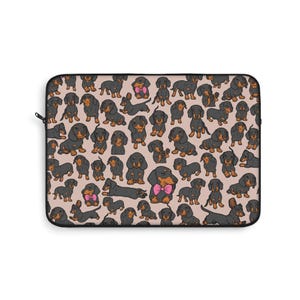 May include: Laptop sleeve featuring a repeating pattern of cartoon dachshunds in various poses. The dogs are black and tan with some wearing pink bows. The background is a light tan color. The sleeve has a black zipper.