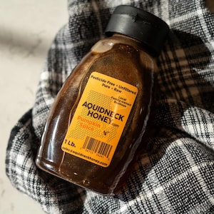 May include: A clear plastic bottle of AQUIDNECK HONEY Pumpkin Spice, with a black cap. The label is orange with black text, stating "Pesticide Free, Unfiltered, Pure, Raw." The bottle contains 1 lb of honey and rests on a black and white checkered cloth.