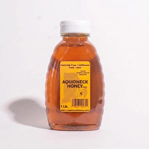 May include: A clear plastic bottle of honey with a white cap. The label reads "AQUIDNECK HONEY.com" and "1 Lb." The honey is amber-colored and the bottle has a ridged design.