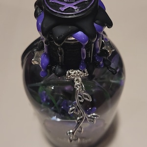 May include: A glass bottle with a black and purple wax seal featuring a skull and crossbones. A silver charm hangs from the bottle's neck.
