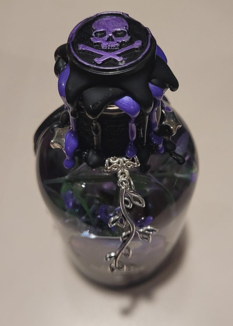 May include: A glass bottle with a black and purple wax seal featuring a skull and crossbones. A silver charm hangs from the bottle's neck.