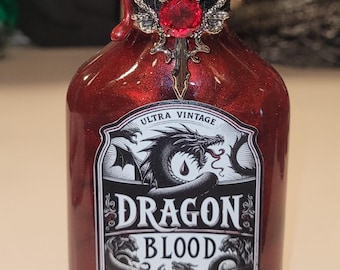 May include: A red glass bottle with a decorative silver stopper and a red gem. The label reads "DRAGON BLOOD" with a dragon illustration. The bottle has a vintage aesthetic.