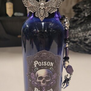 May include: A dark blue glass bottle with a black and purple wax seal. The bottle has a silver charm of a moth with a skull on its body. The bottle has a label that reads "Poison Potions".
