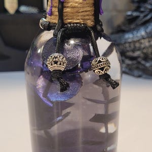 May include: A small glass bottle with a purple wax seal and black twine. The bottle is filled with a purple liquid and contains small silver beads.