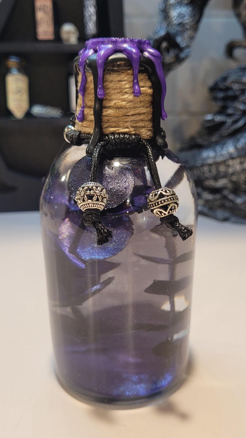 May include: A small glass bottle with a purple wax seal and black twine. The bottle is filled with a purple liquid and contains small silver beads.