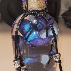 May include: A glass bottle with a purple wax seal and a black cord tied around the neck. The bottle is filled with a blue and black glittery liquid and contains a blue and black flower.