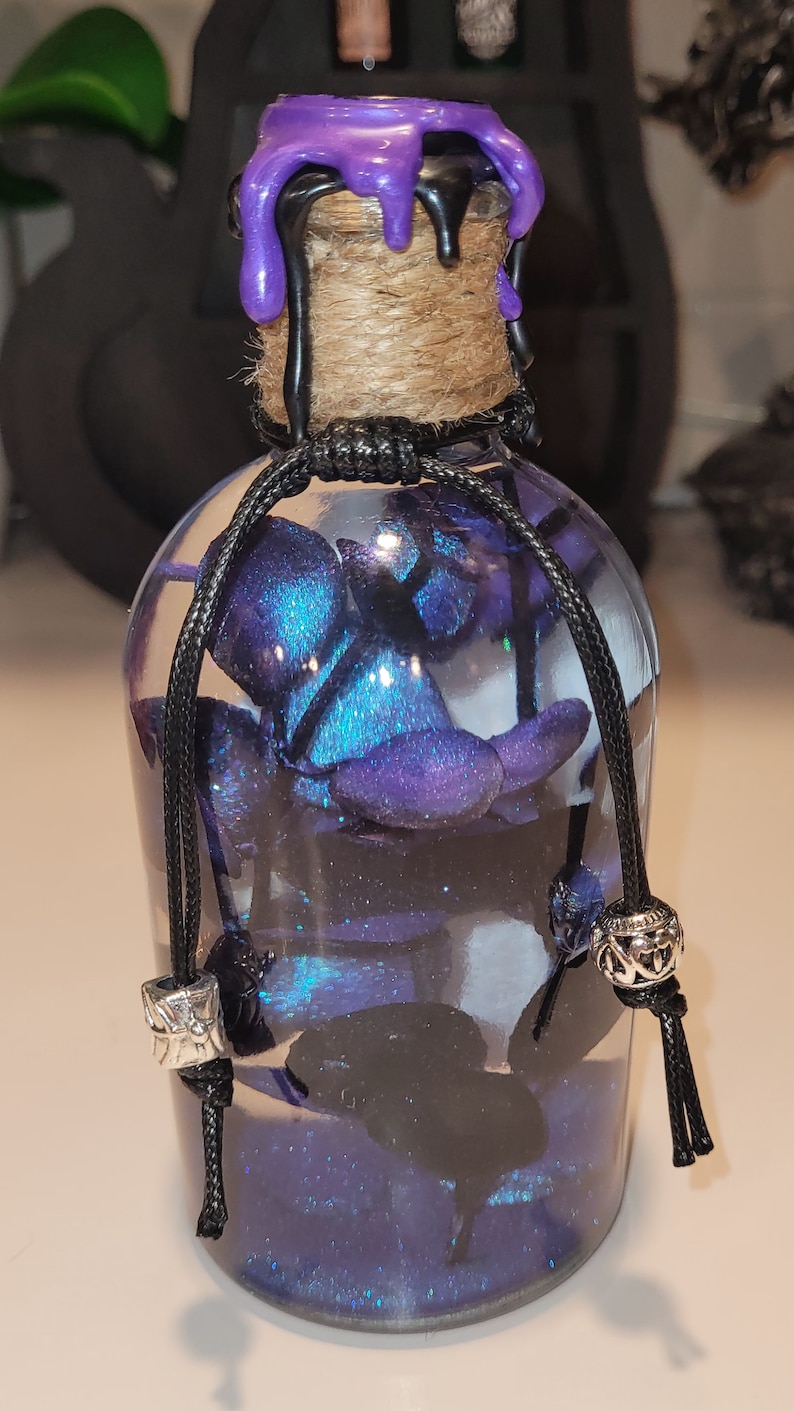May include: A glass bottle with a purple wax seal and a black cord tied around the neck. The bottle is filled with a blue and black glittery liquid and contains a blue and black flower.