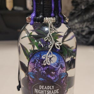 May include: A glass bottle with a black and purple wax seal, a silver charm, and a sticker that says "Deadly Nightshade". The bottle is filled with a clear liquid and green foliage.
