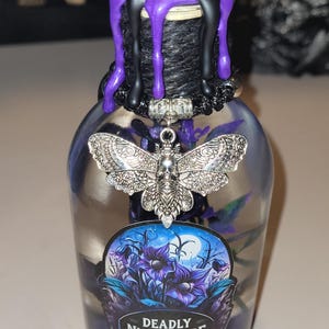 May include: A clear glass bottle with a black and purple wax seal. The bottle contains a purple liquid and a silver charm of a moth with a skull. The label on the bottle reads "Deadly Nightshade".
