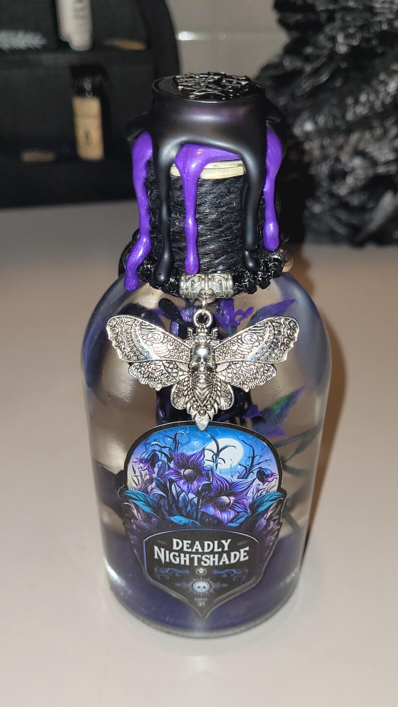 May include: A clear glass bottle with a black and purple wax seal. The bottle contains a purple liquid and a silver charm of a moth with a skull. The label on the bottle reads "Deadly Nightshade".