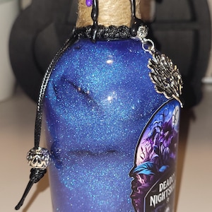 May include: A small glass bottle with a blue glitter liquid and a black and purple wax seal. The bottle has a black cord with a silver charm and a label that reads "Deadly Nightshade".