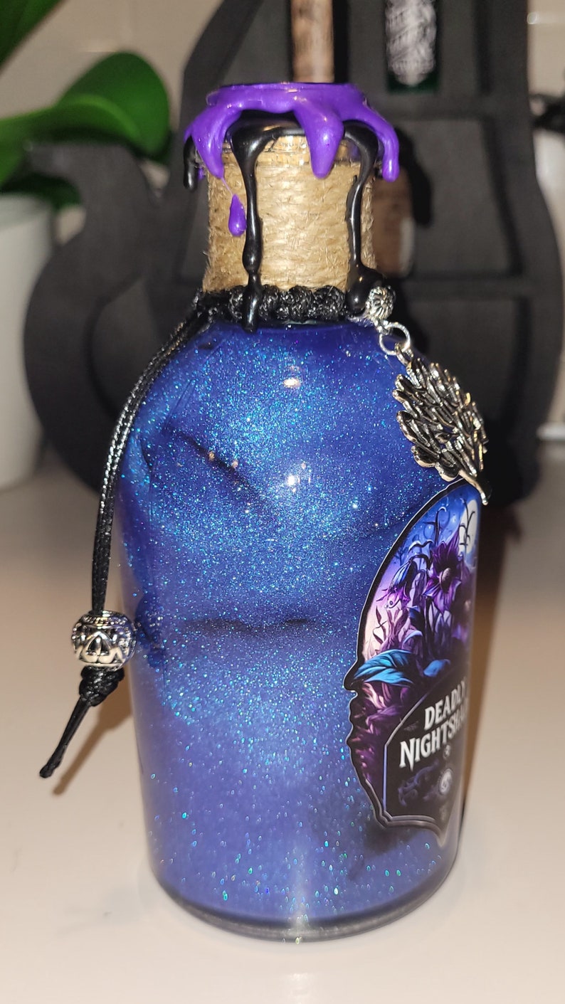 May include: A small glass bottle with a blue glitter liquid and a black and purple wax seal. The bottle has a black cord with a silver charm and a label that reads "Deadly Nightshade".